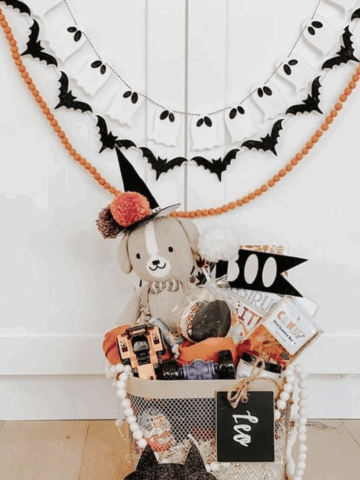 Neutral Halloween boo basket with a plush dog toy, festive treats, and black cat mask, displayed under ghost and bat garland decor