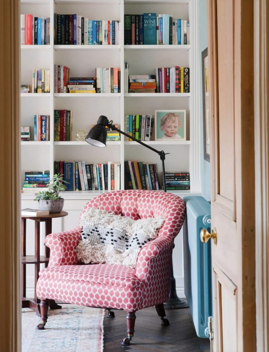 Reading nook with a pink patterned armchair, textured throw pillow, floor lamp, and white bookshelves filled with colorful books