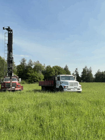 Front view of drilling trucks on a rural property preparing to drill a water well in a grassy field with forest backdrop.