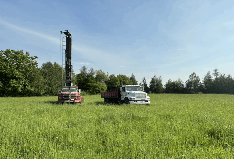 Front view of drilling trucks on a rural property preparing to drill a water well in a grassy field with forest backdrop.