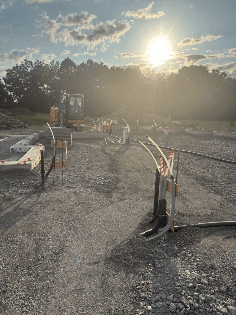 Wide view of underground electrical and utility conduits installed before slab pour, with excavator and early morning sunlight in the background.
