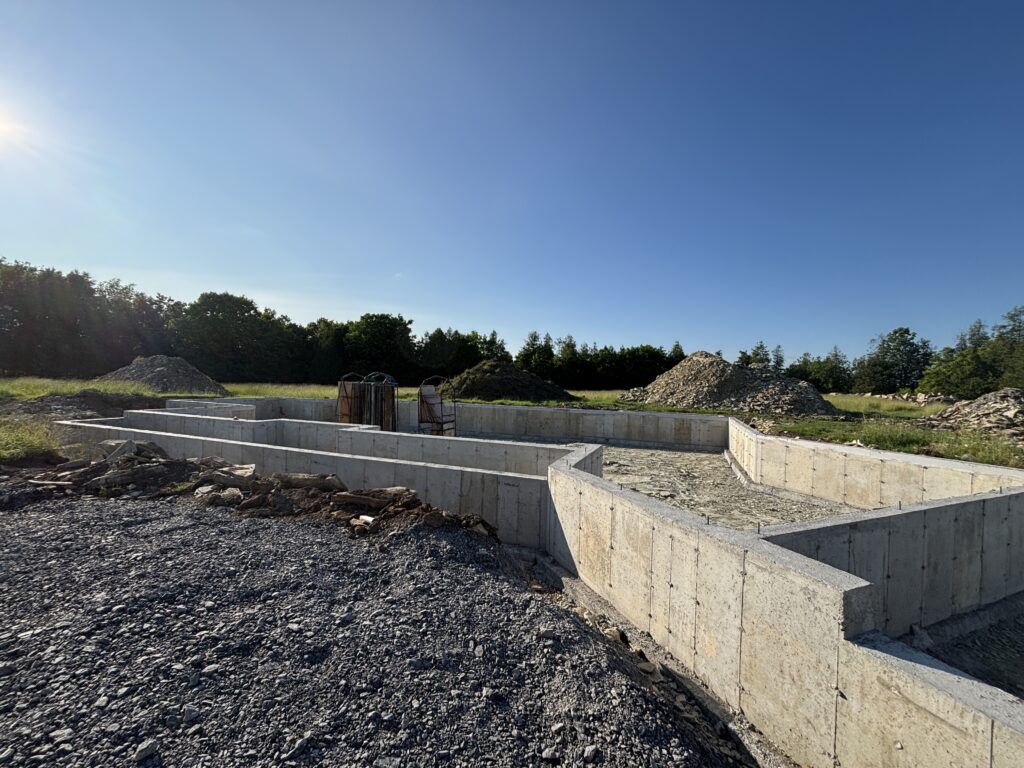 Completed concrete foundation walls under blue sky, surrounded by piles of earth and building materials.
