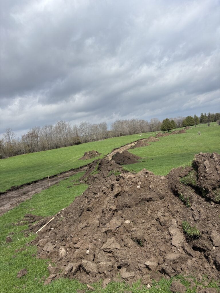 Piles of topsoil line a curved section of land during rural driveway excavation on a green field.