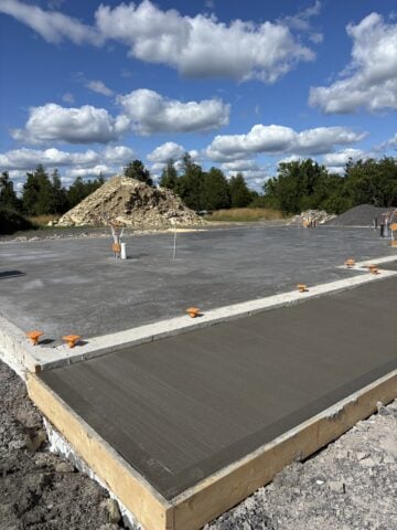 Slab-on-grade foundation and freshly poured concrete entry pad during the early phase of building our home, with utility rough-ins and gravel piles visible