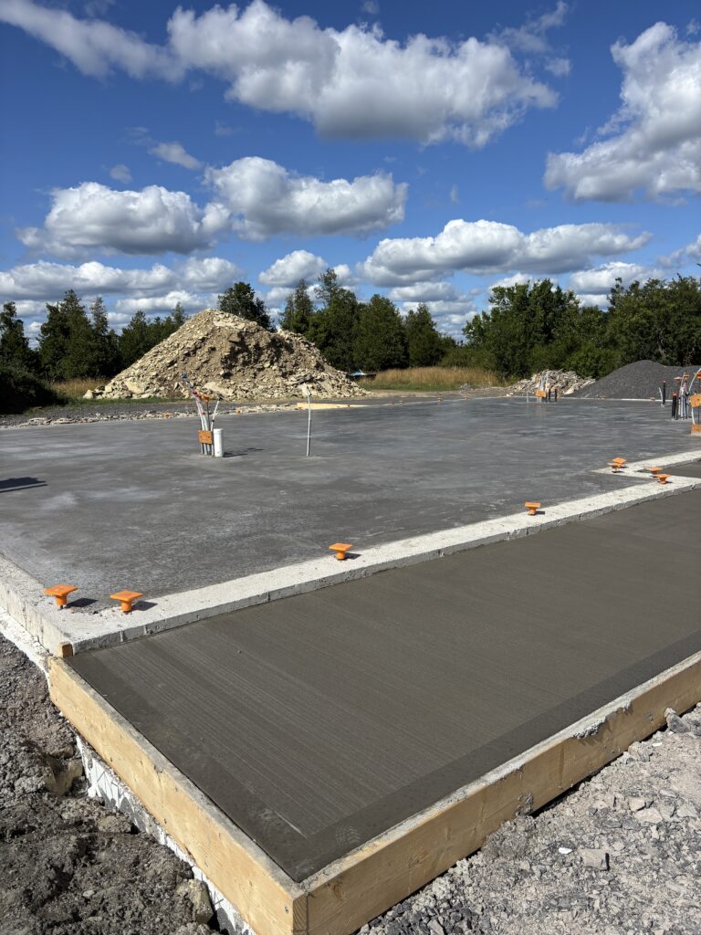 Slab-on-grade foundation and freshly poured concrete entry pad during the early phase of building our home, with utility rough-ins and gravel piles visible