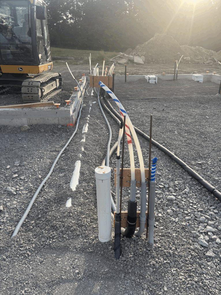 Close-up of multiple electrical, water, and conduit lines organized and staked in gravel during foundation prep before concrete pour.