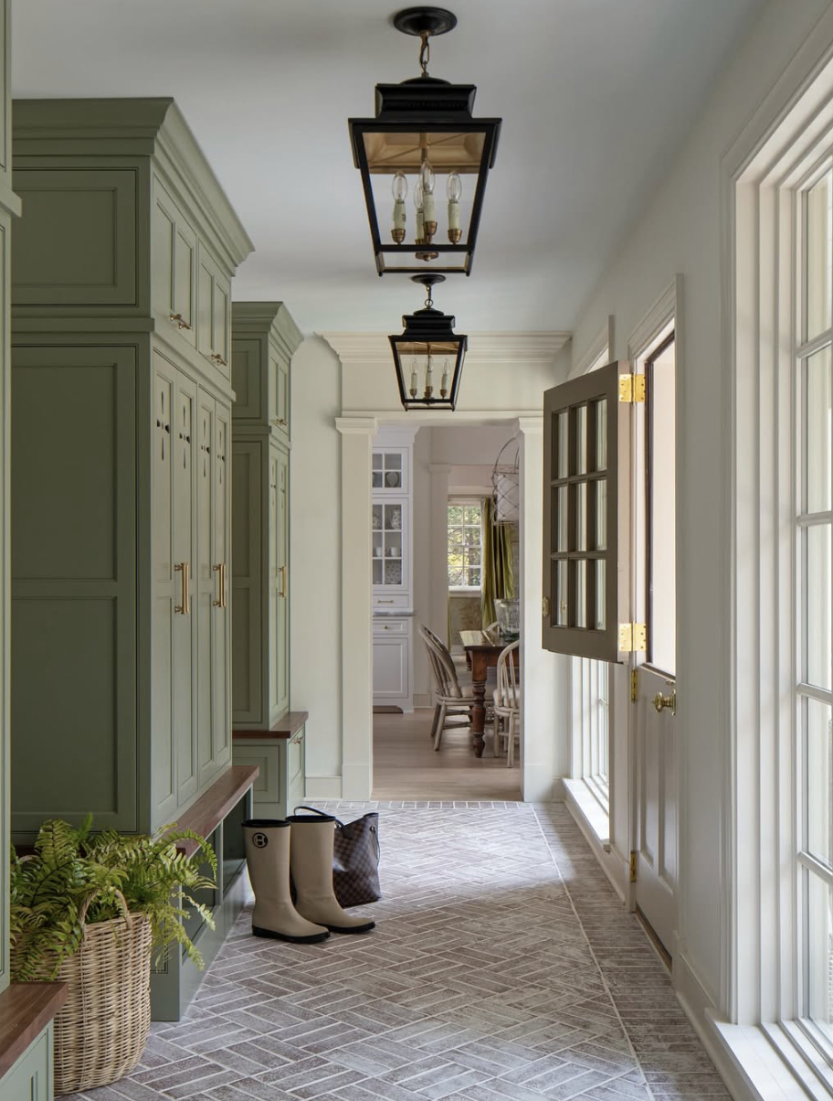 Mudroom with deep sage green built-in cabinets, wood bench seating, and herringbone tile flooring.