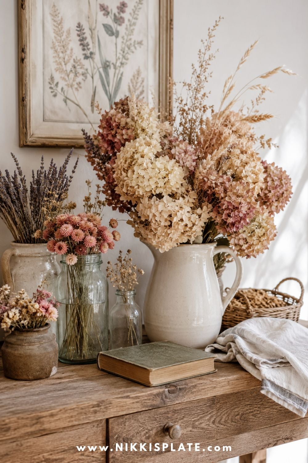Dried hydrangeas, lavender, wheat and strawflowers arranged in vintage jars and stoneware crocks on a warm wood console.