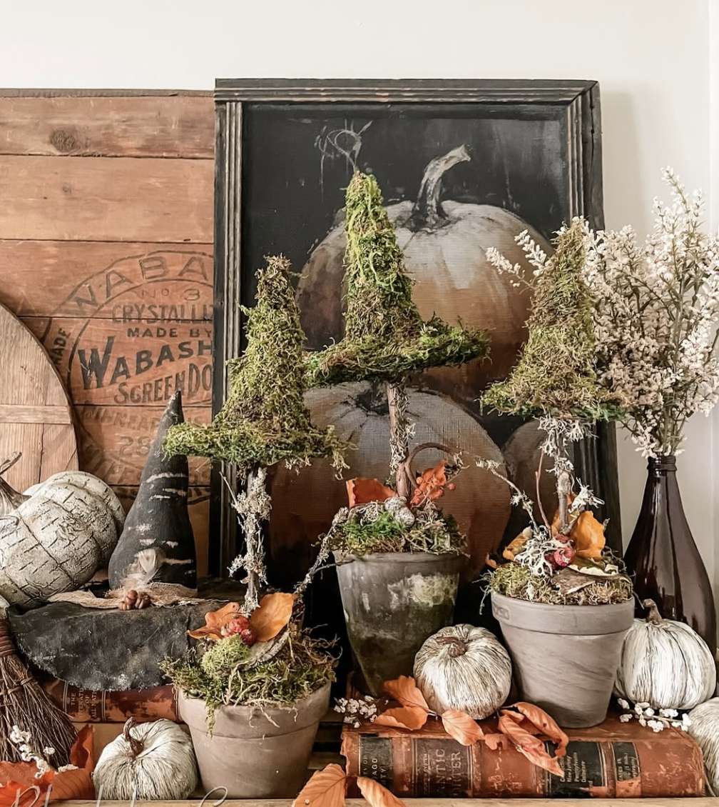 Rustic fall display featuring mossy topiaries, terracotta pots, muted pumpkins, dried leaves, and vintage books in warm, earthy tones of brown, clay, olive, and ivory.
