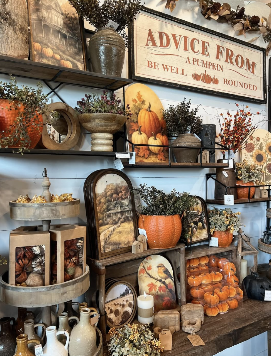 Fall-themed wall display featuring dark wood shelves, vintage autumn artwork, ceramic vases, mini pumpkins, and warm-toned florals in a cozy retail setting.