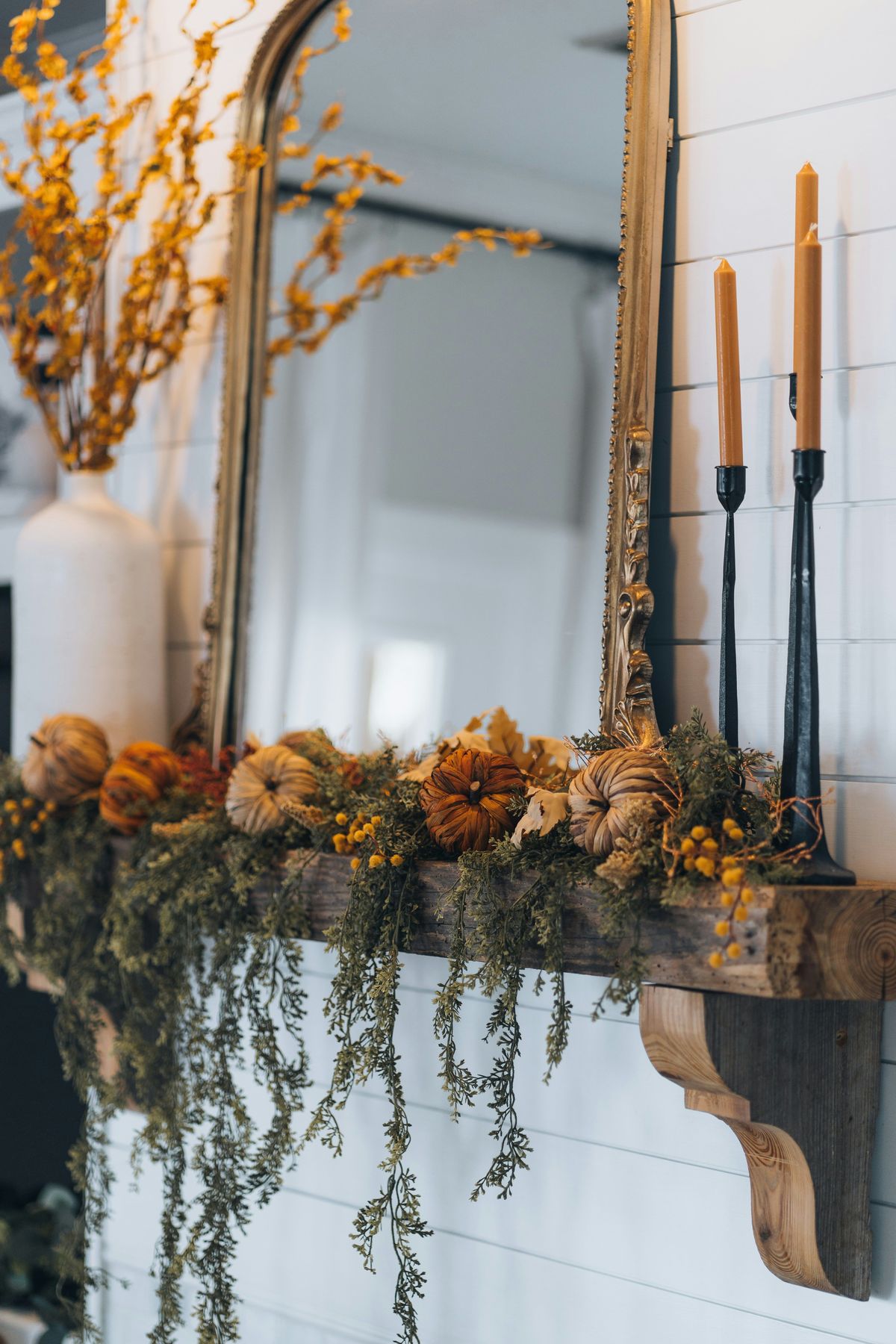 Rustic fall mantel decorated with dried pumpkins, yellow florals, cedar garland, and taper candles for a nature-inspired seasonal look.