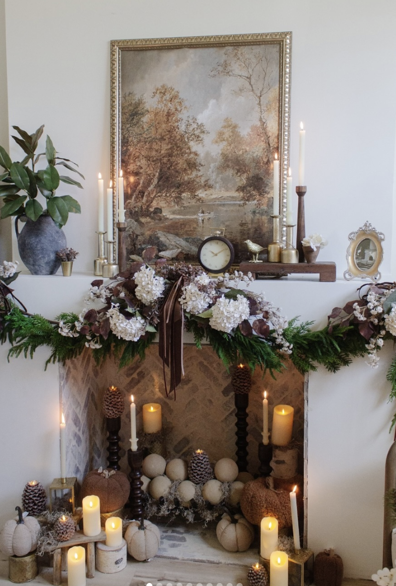 Sophisticated fall fireplace display with sculptural candleholders, gold-toned accents, layered garland, and classic artwork in a warm, vintage-inspired setting.