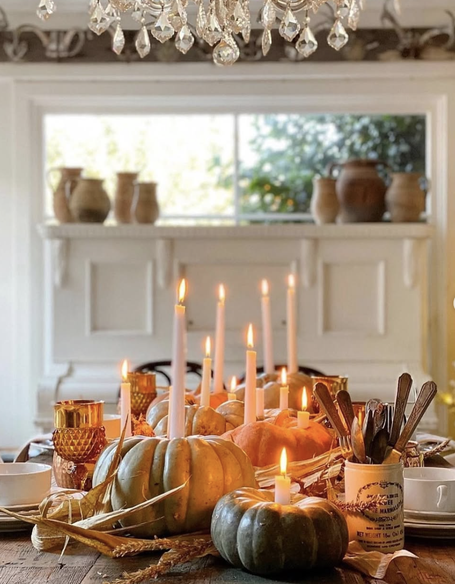 Elegant fall dining table centerpiece featuring heirloom pumpkins, tapered candles, amber glassware, and vintage-style flatware under a crystal chandelier.