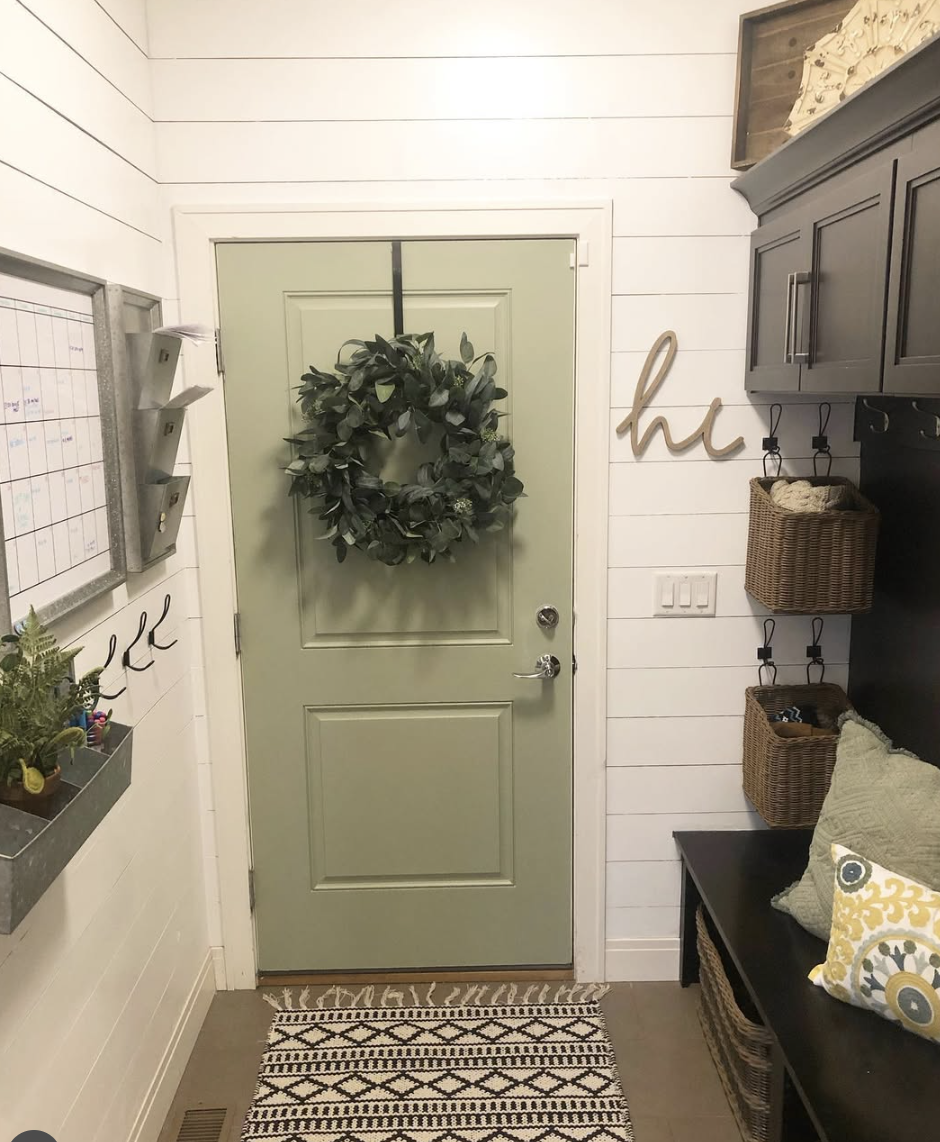 Soft green entry door with a wreath, shiplap walls, farmhouse-style storage bench, and woven baskets.