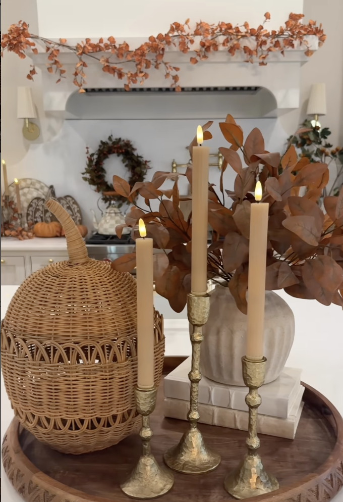 Fall kitchen vignette featuring brass candle holders, faux autumn leaves, a wicker pumpkin, and neutral stacked books on a wooden tray, with orange garland and wreaths in the background.