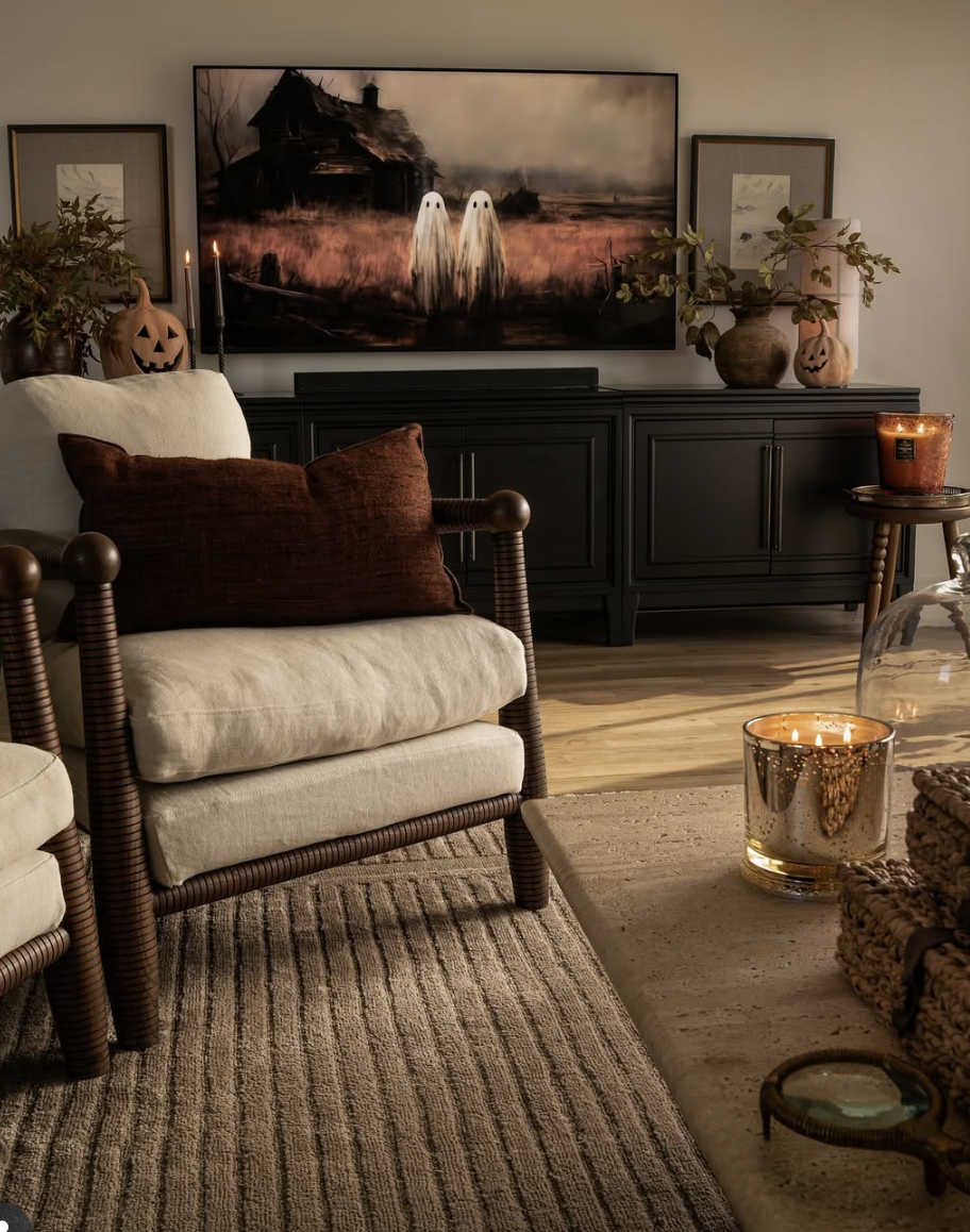 Warm and moody fall living room with neutral furniture, brown accents, black media console, jack-o’-lanterns, candlelight, and Halloween art displayed on the TV screen.