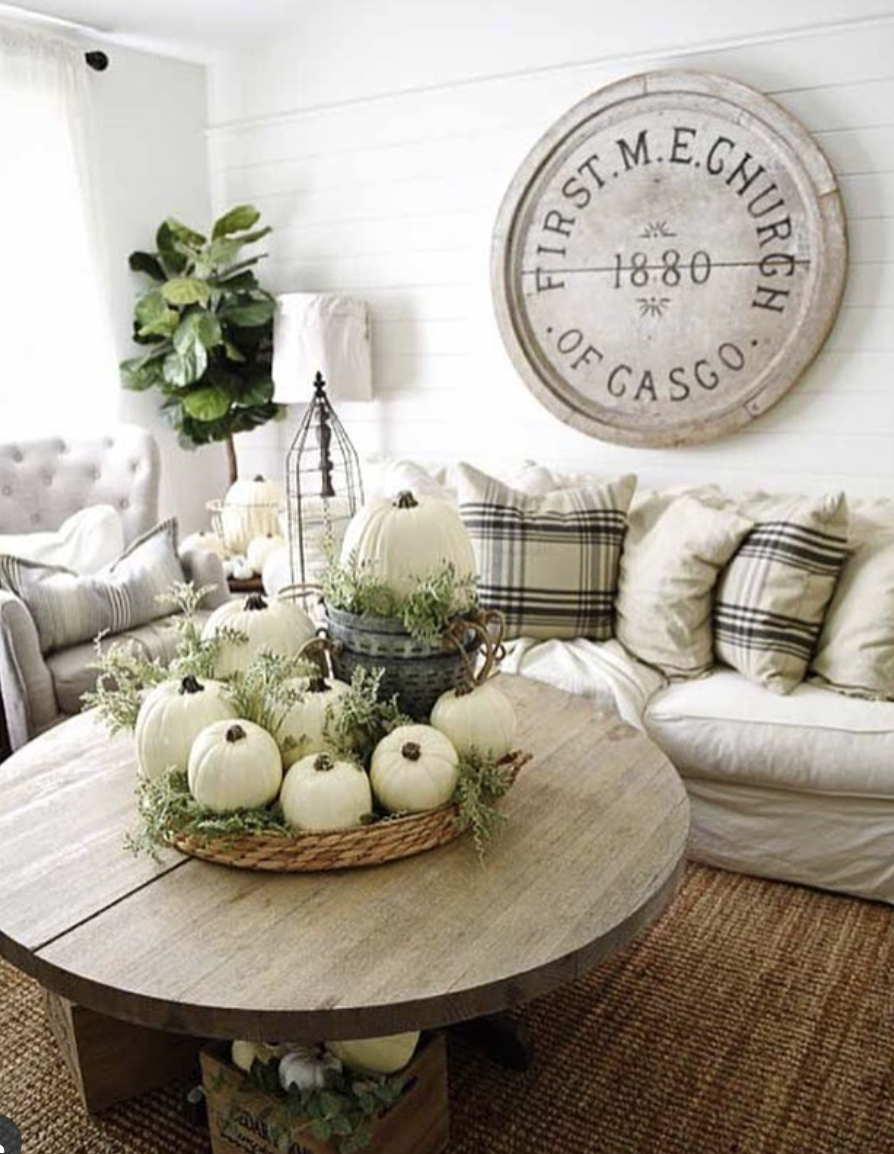 Farmhouse-style fall living room featuring oversized plaid throw pillows, white pumpkins with greenery on a round wood coffee table, and a rustic church sign wall decor.