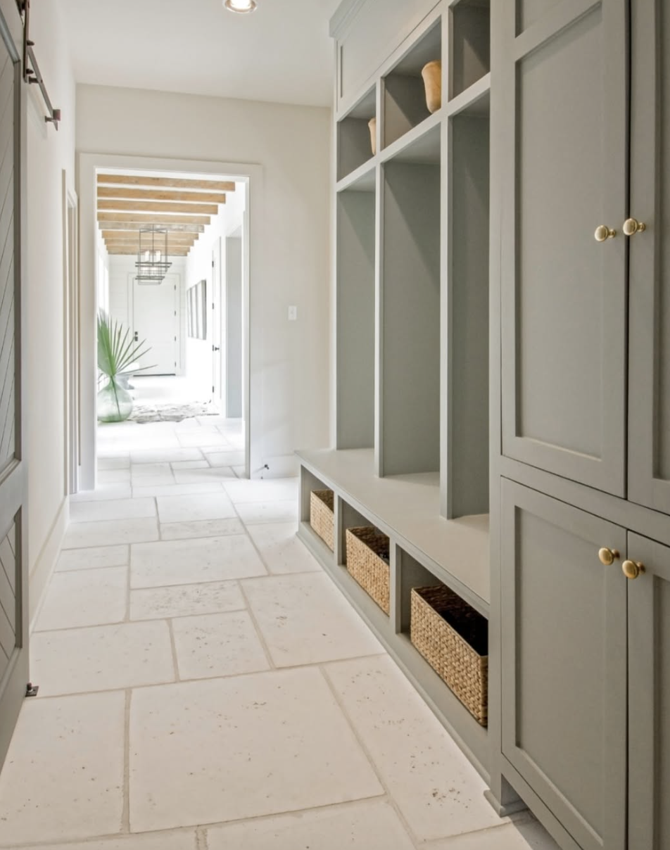 Mudroom with sage green built-in lockers, open cubbies with woven baskets, and large stone tile flooring.