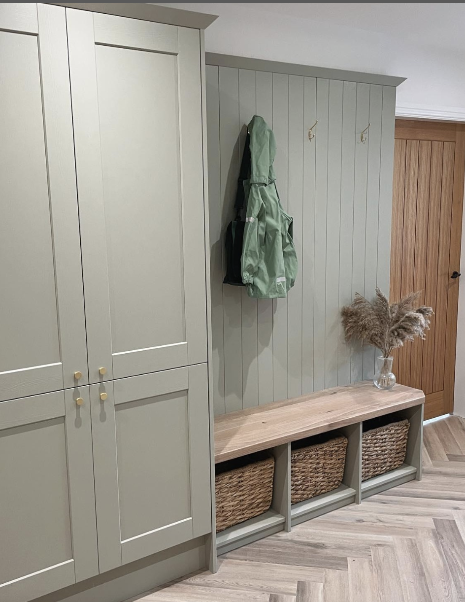 Sage green mudroom featuring built-in cabinets, a wood-topped bench with basket storage, and vertical panelled wall with coat hooks.