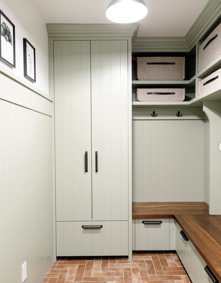 Sage green mudroom with vertical paneling, built-in storage cabinets, brick herringbone flooring, and wood bench seating.