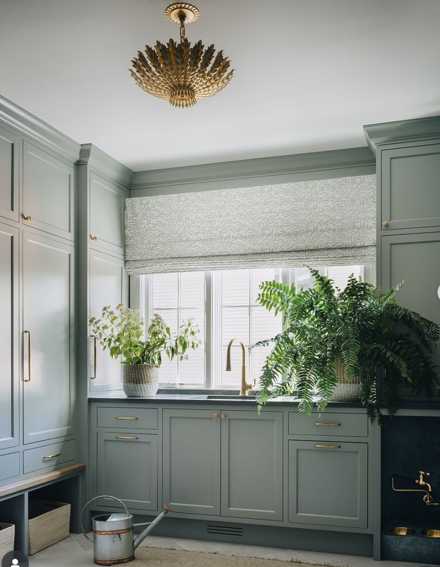Sage green mudroom with shaker cabinets, built-in sink, brass hardware, and potted plants by a large window.