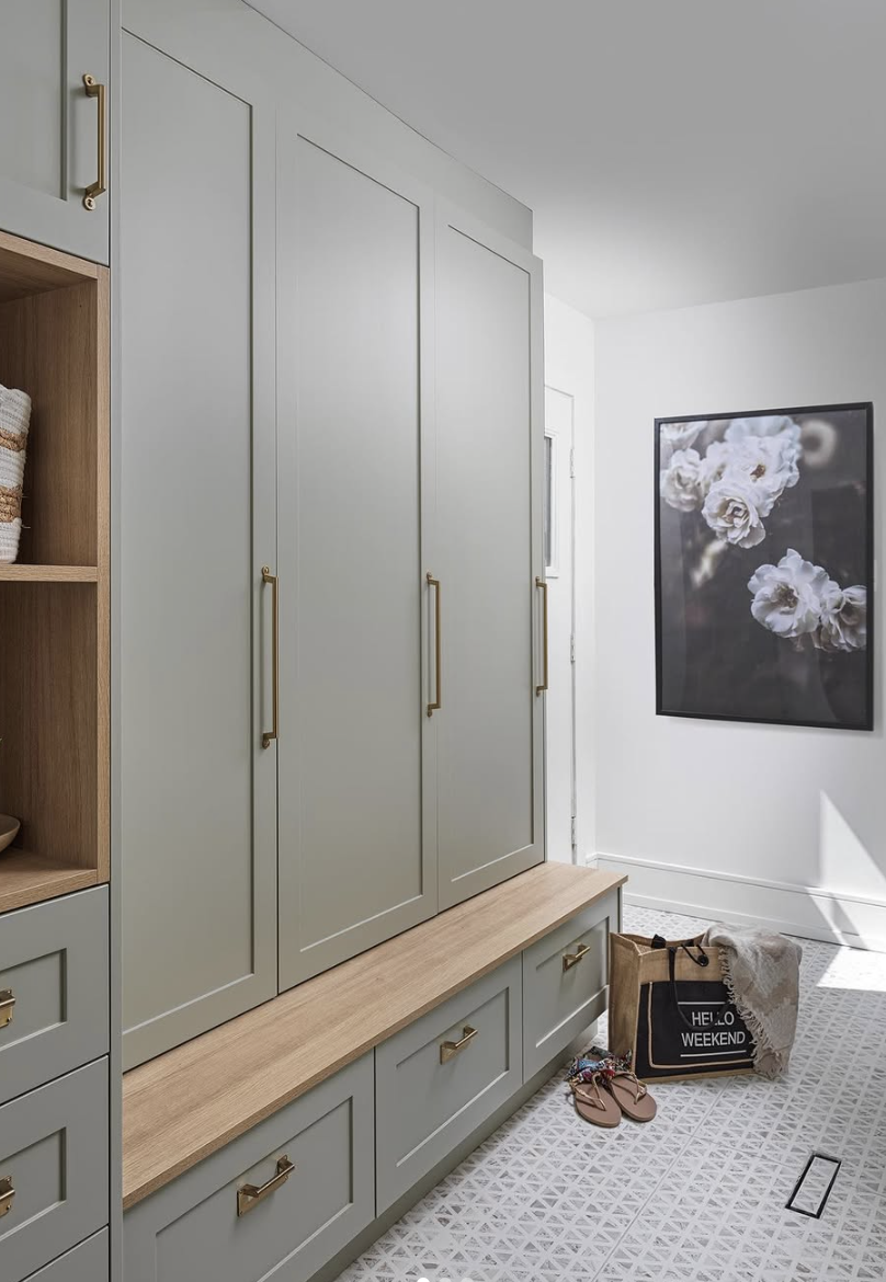 Sage green mudroom with built-in cabinetry, gold hardware, wood bench seating, and patterned tile flooring.