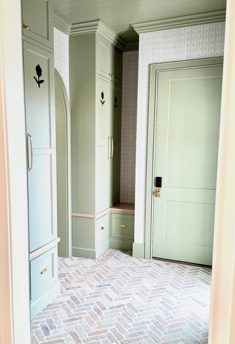 Sage green mudroom with shaker cabinetry, brass hardware, built-in seating, and herringbone brick flooring.