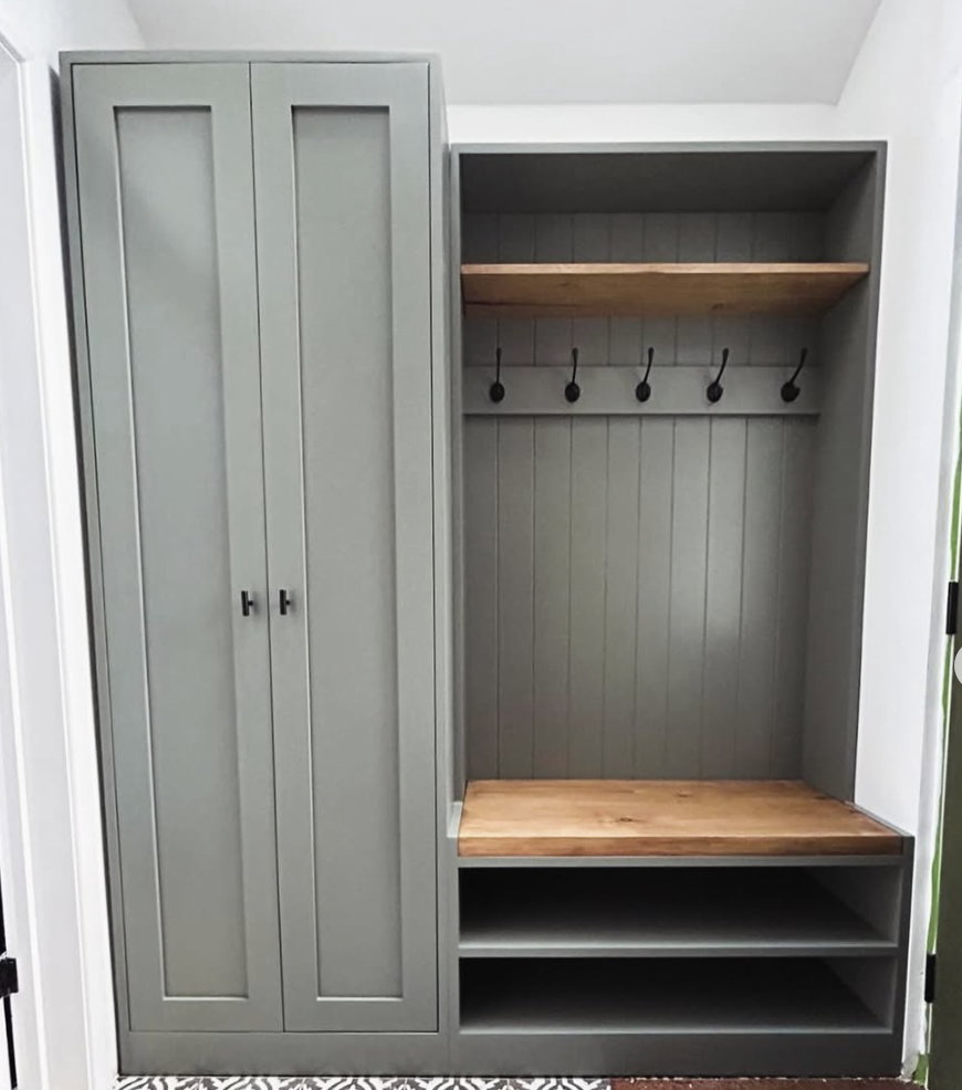 Minimalist sage green mudroom with shaker-style cabinet, open bench seating, wood shelf, and black hooks.