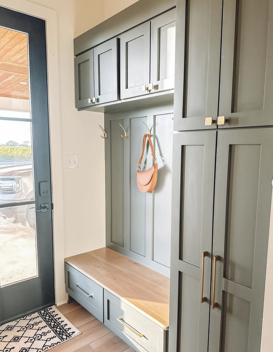 Sage green mudroom with shaker-style cabinetry, upper storage cabinets, wood bench seating, and gold hardware.