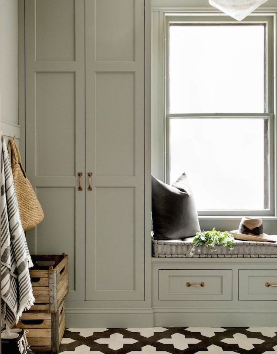 Sage green mudroom featuring tall shaker-style cabinets, built-in window bench with drawers, and patterned floor tiles.