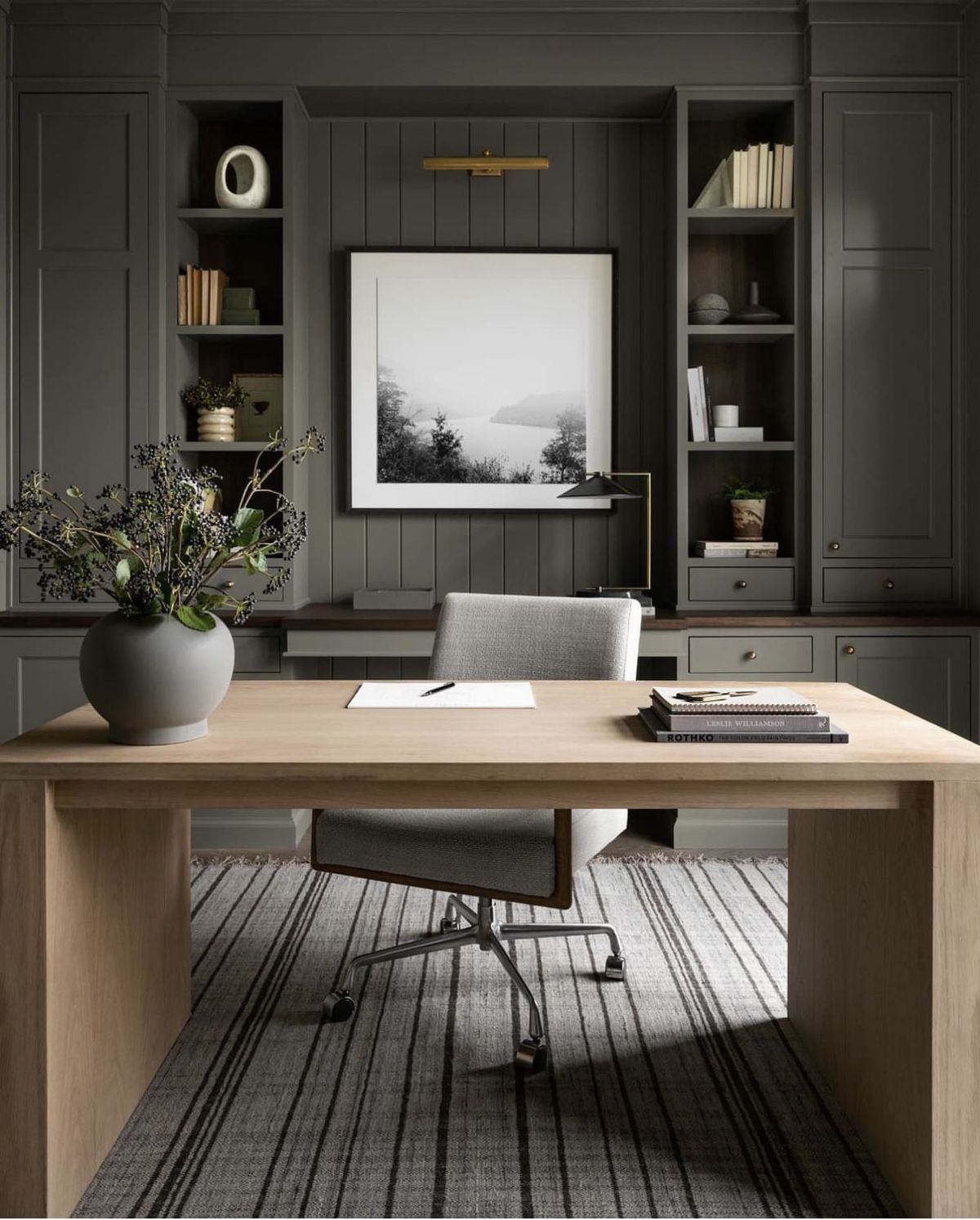 Studio McGee office featuring gray built-in cabinetry, a light wood desk, striped rug, and a large black-and-white framed artwork.