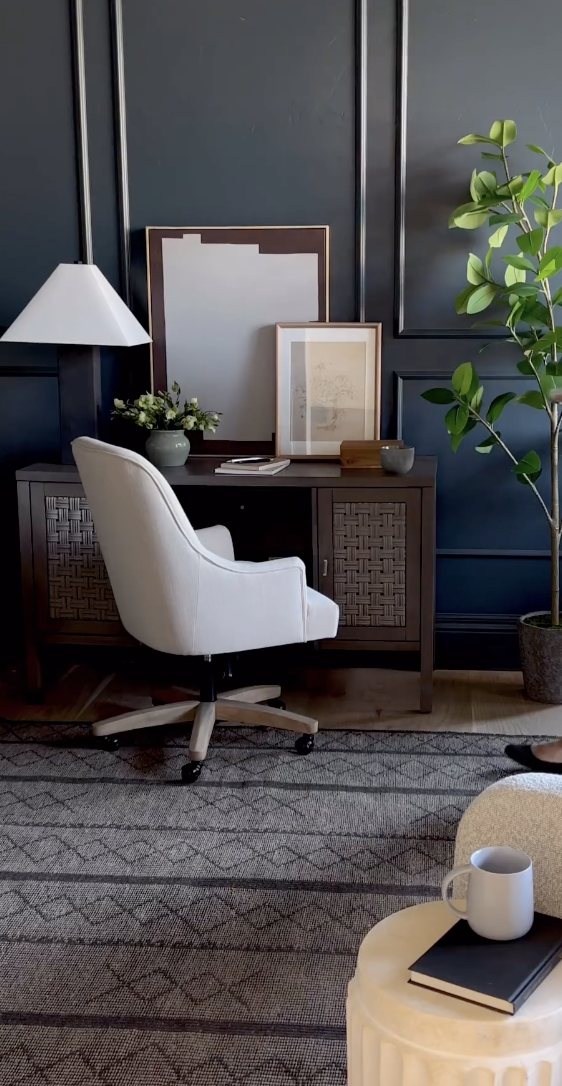 Studio McGee office featuring navy paneled walls, a dark wood desk, white upholstered office chair, and greenery accents.