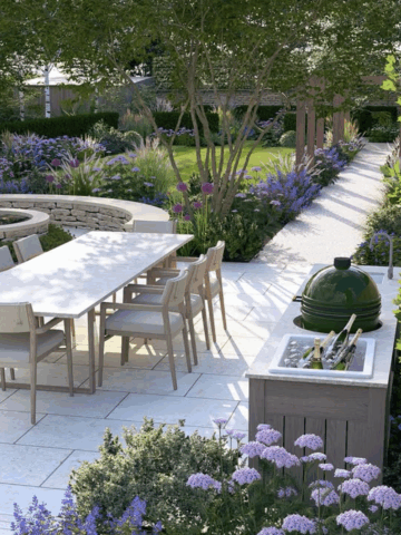 A beautifully landscaped backyard with a white stone patio, outdoor dining table, lush purple flowers, and a shaded garden path — perfect inspiration for a backyard refresh.