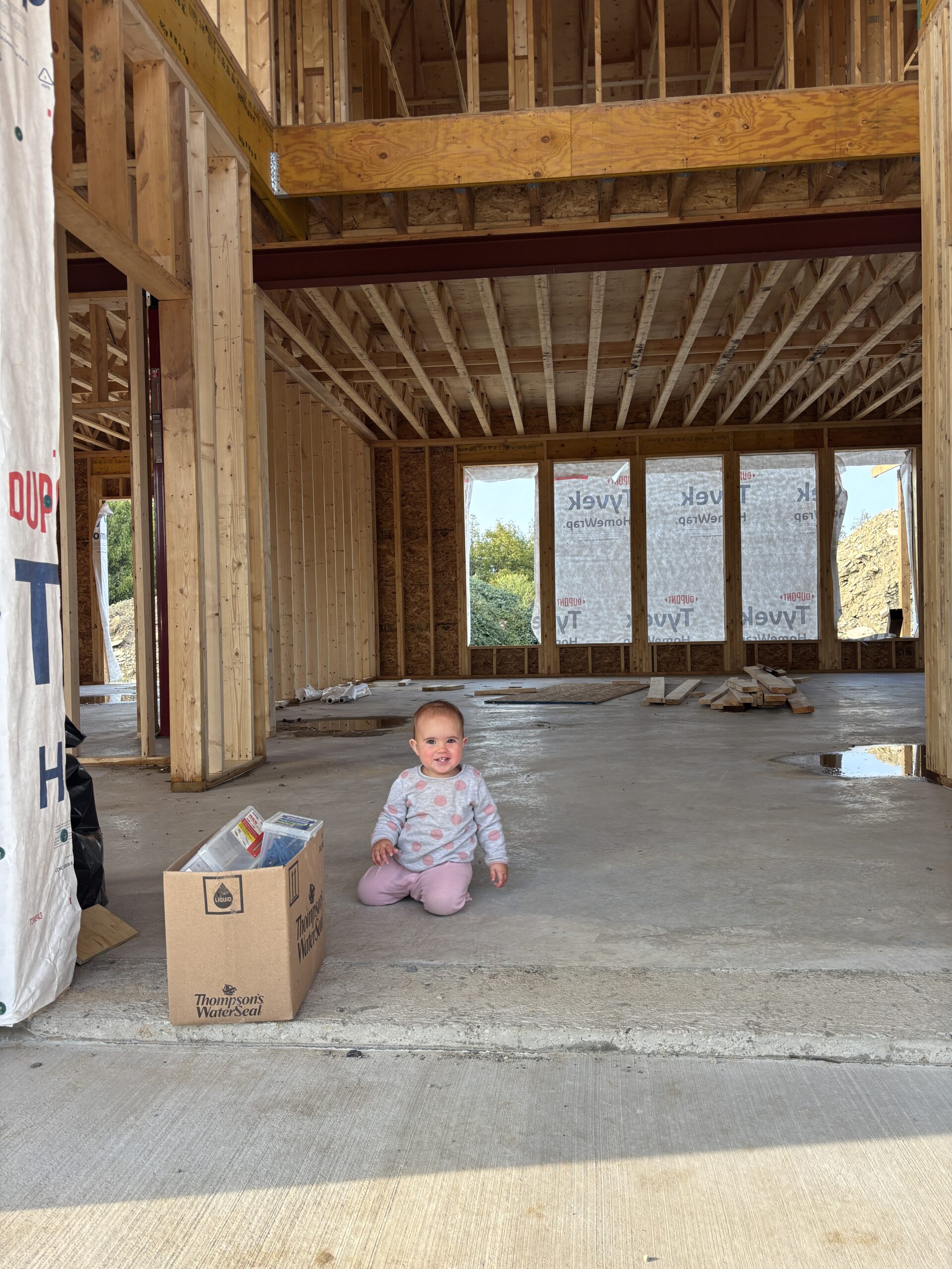Baby sitting inside a framed modern farmhouse under construction, surrounded by wooden beams and unfinished walls during the building process.