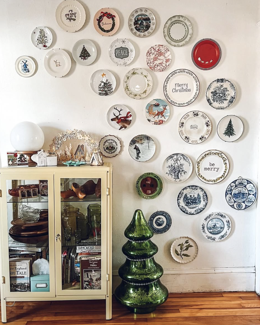 Gallery-style Christmas plate wall with assorted holiday plates, woodland scenes, and festive sayings above a vintage cabinet and green glass tree.
