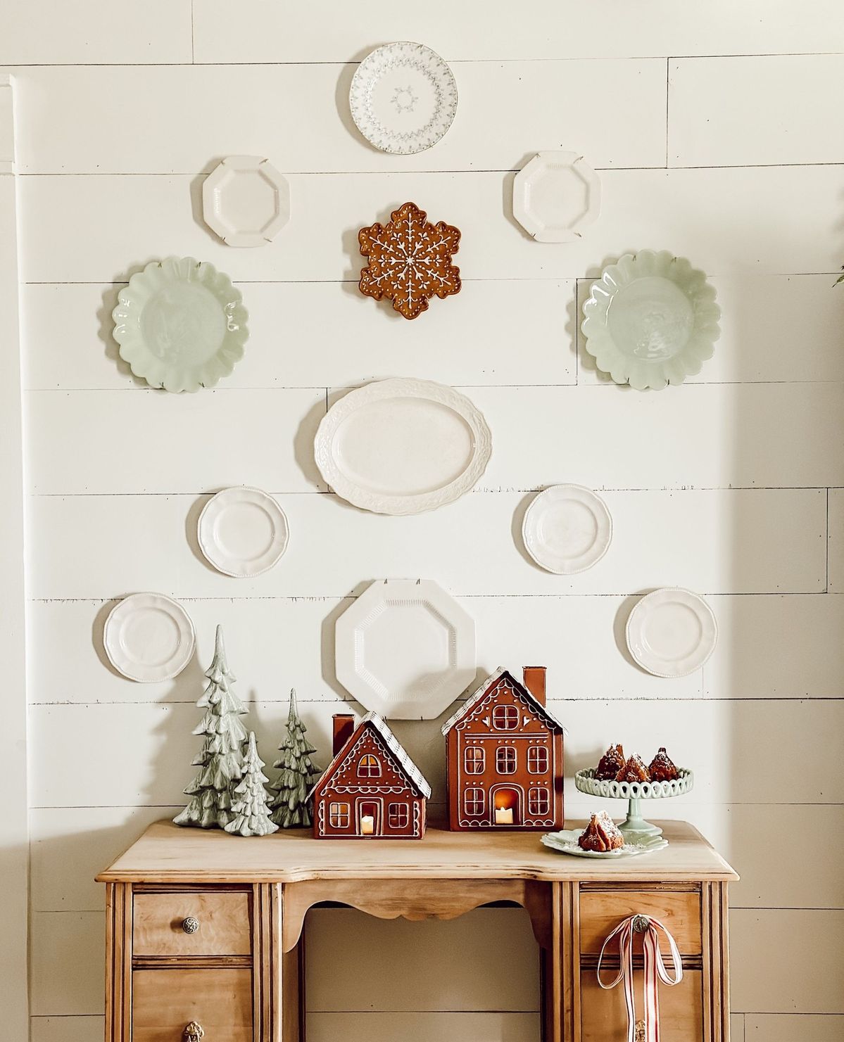 Christmas plate wall with neutral plates and a single gingerbread snowflake plate, styled with ceramic trees and gingerbread houses on a wood desk.