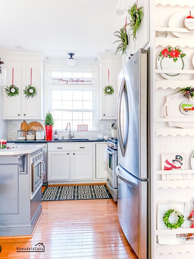 White kitchen with built-in plate rack featuring Christmas plates and mini wreaths hanging on cabinets for a festive holiday look.