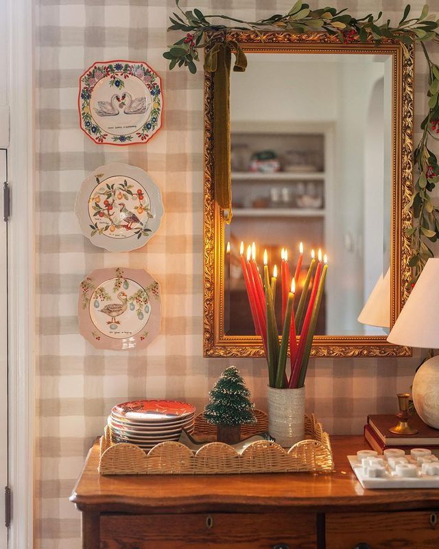 Christmas plate wall with three decorative holiday plates beside a gold mirror, styled with greenery garland and tall red candles.
