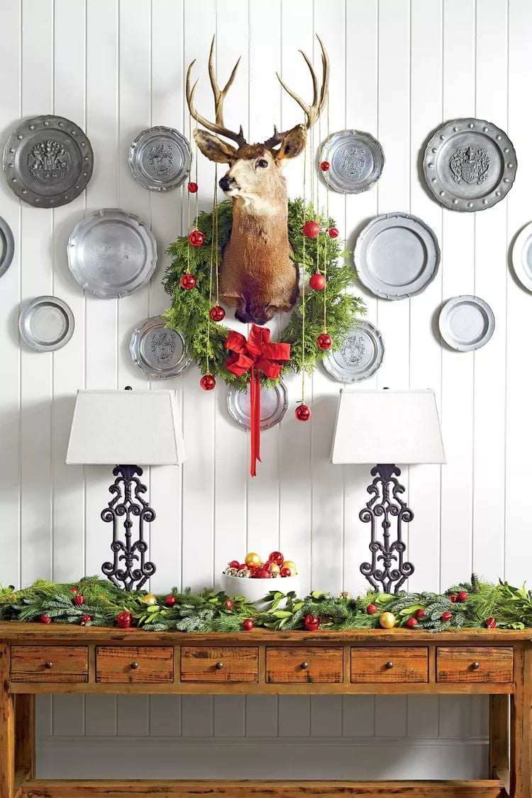 Christmas plate wall with vintage pewter plates arranged around a deer mount with greenery wreath and red ornaments, styled above a wood console table.