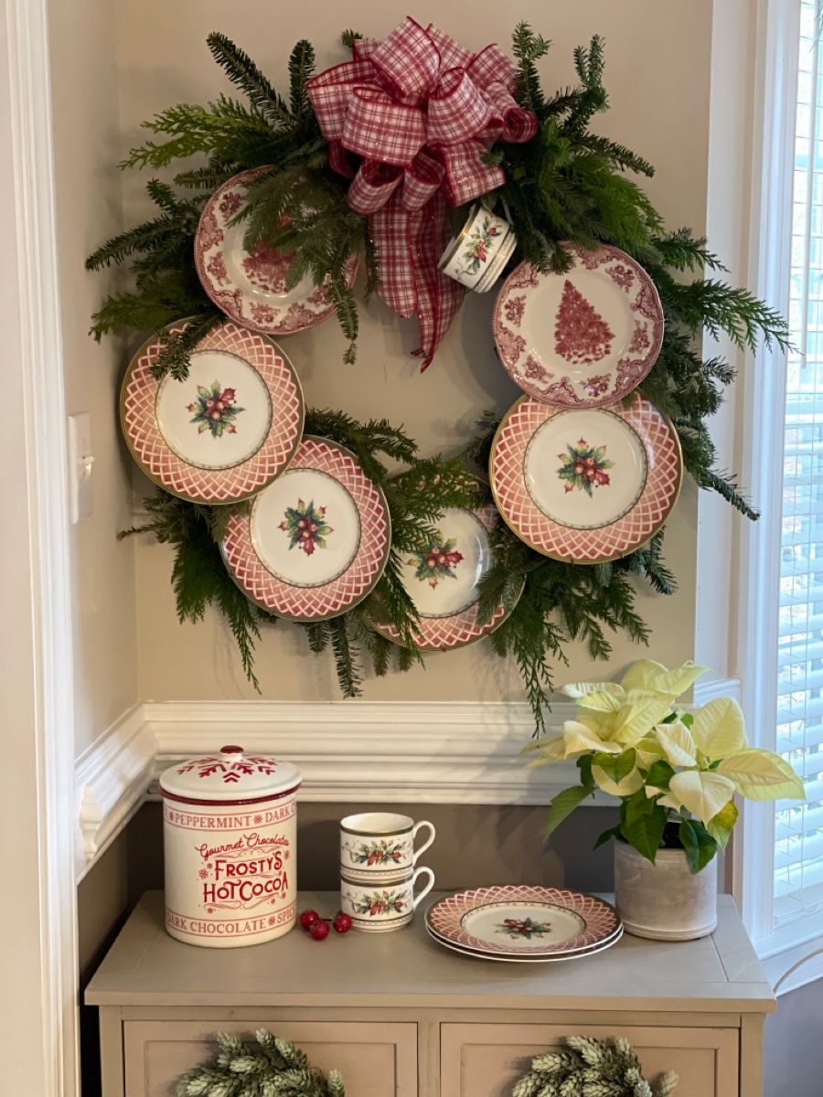 Christmas plate wall wreath with red and white holiday china, plaid bow, and fresh greenery over a hot cocoa station.