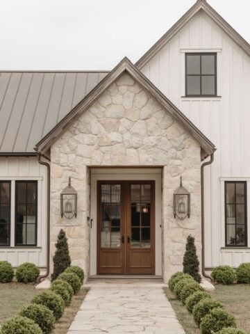 Charming stone farmhouse entryway with double wood front doors, black windows, and boxwood-lined path creating a warm and welcoming first impression.
