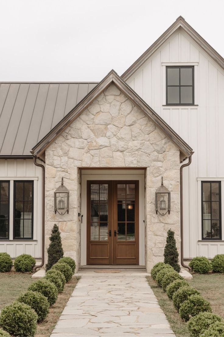 Charming stone farmhouse entryway with double wood front doors, black windows, and boxwood-lined path creating a warm and welcoming first impression.