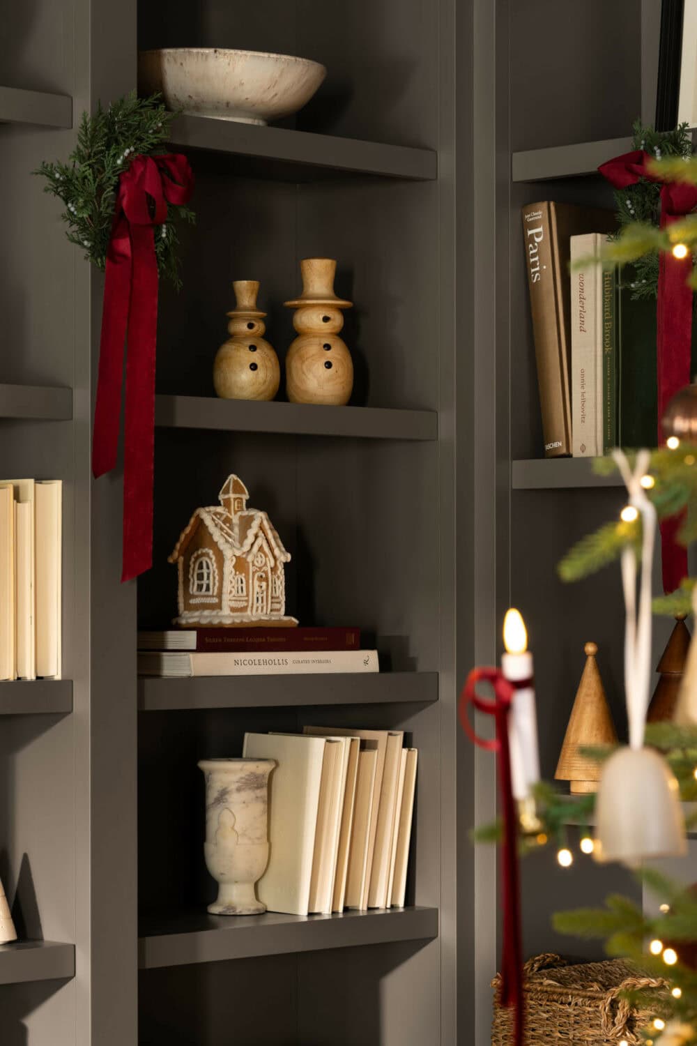 Studio McGee styled bookshelf with Christmas decor including wooden snowmen, gingerbread house, greenery, and red ribbon accents.