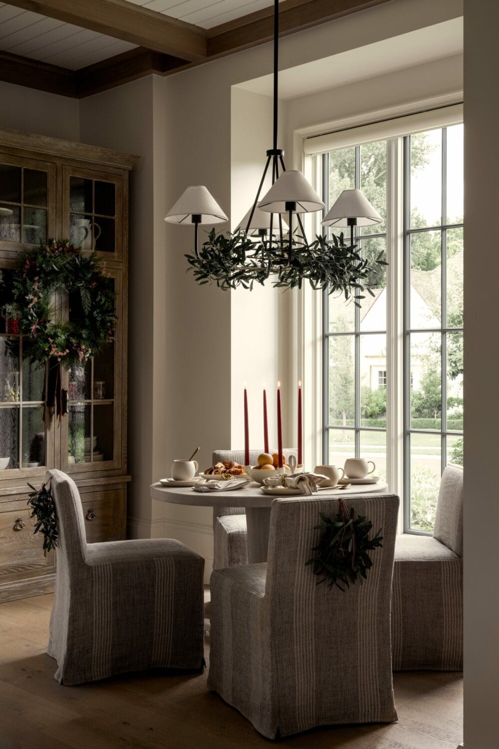Studio McGee breakfast nook decorated for Christmas with mini wreaths on chairs, greenery chandelier, and red holiday candles.