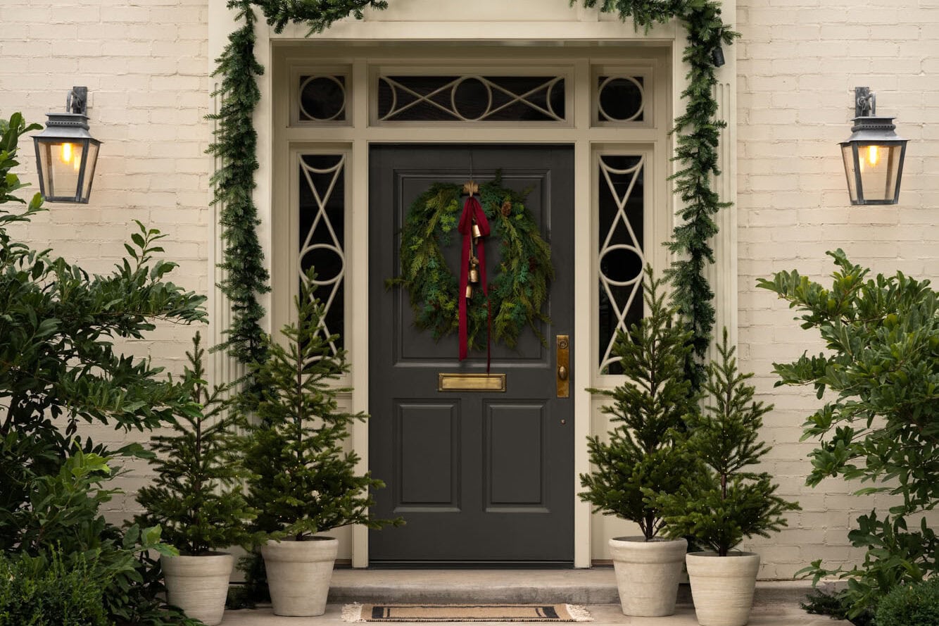 Studio McGee Christmas front door with greenery wreath, red ribbon, potted evergreens, and holiday garland framing the entry.