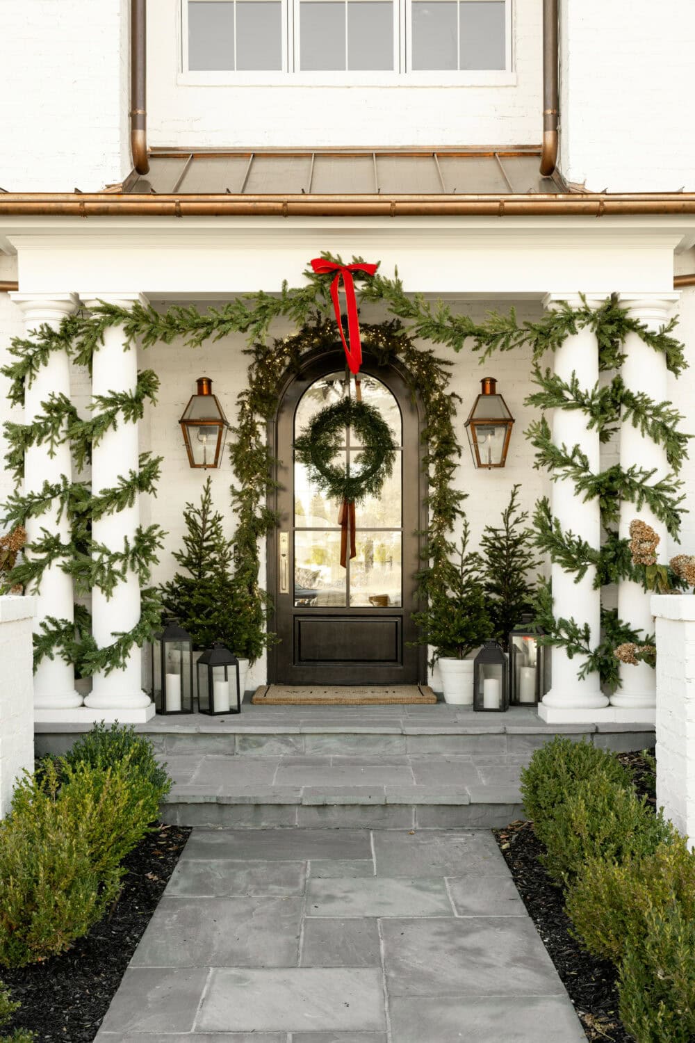 Studio McGee Christmas front porch with greenery garlands on columns, wreath with red ribbon, potted evergreens, and lantern accents.
