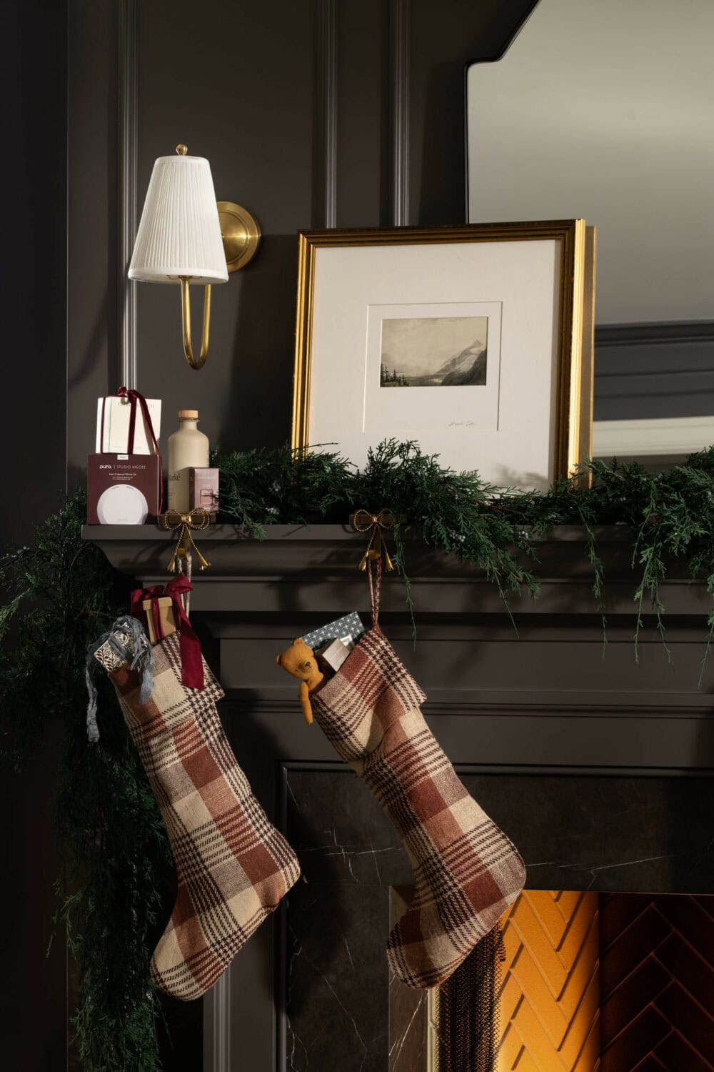 Studio McGee Christmas mantel with plaid stockings, greenery garland, gold accents, and dark-painted fireplace surround.
