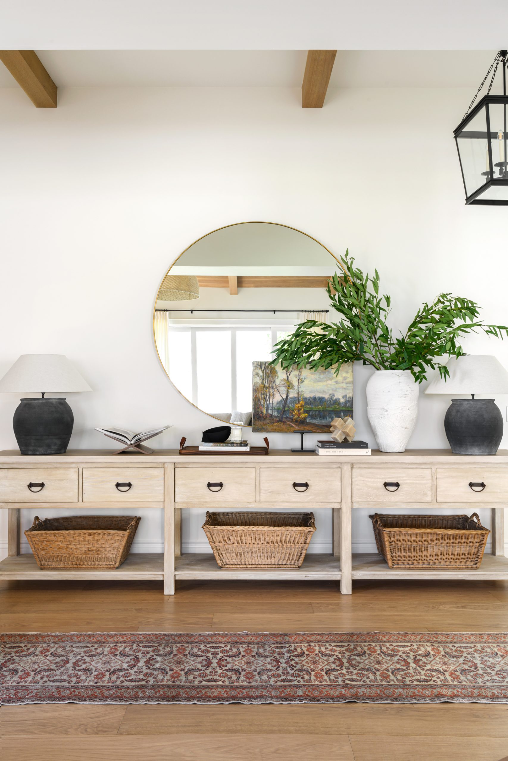 Studio McGee entryway with long light-wood console, round mirror, black lamps, woven baskets, and vintage rug creating a bright modern farmhouse foyer.