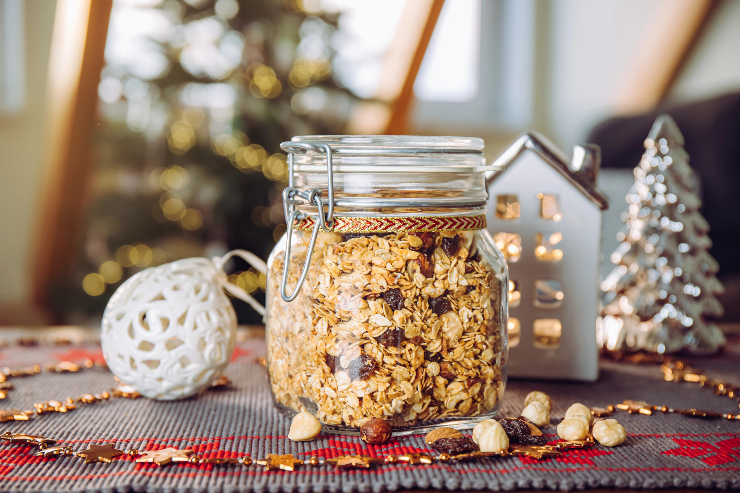Homemade granola in a mason jar with nuts and raisins, decorated with holiday ribbon and Christmas decor in the background.