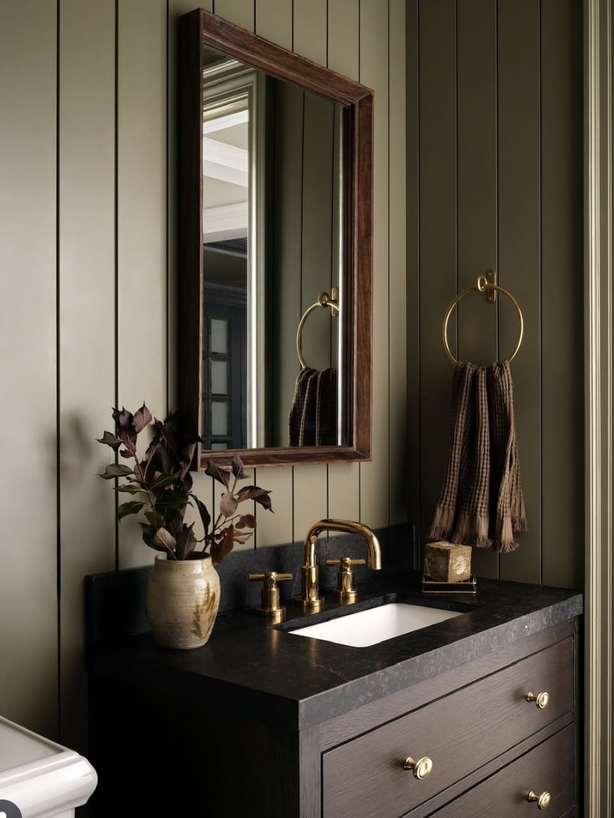 Bathroom with moody green vertical shiplap walls, wood vanity, black countertop, and brass fixtures.